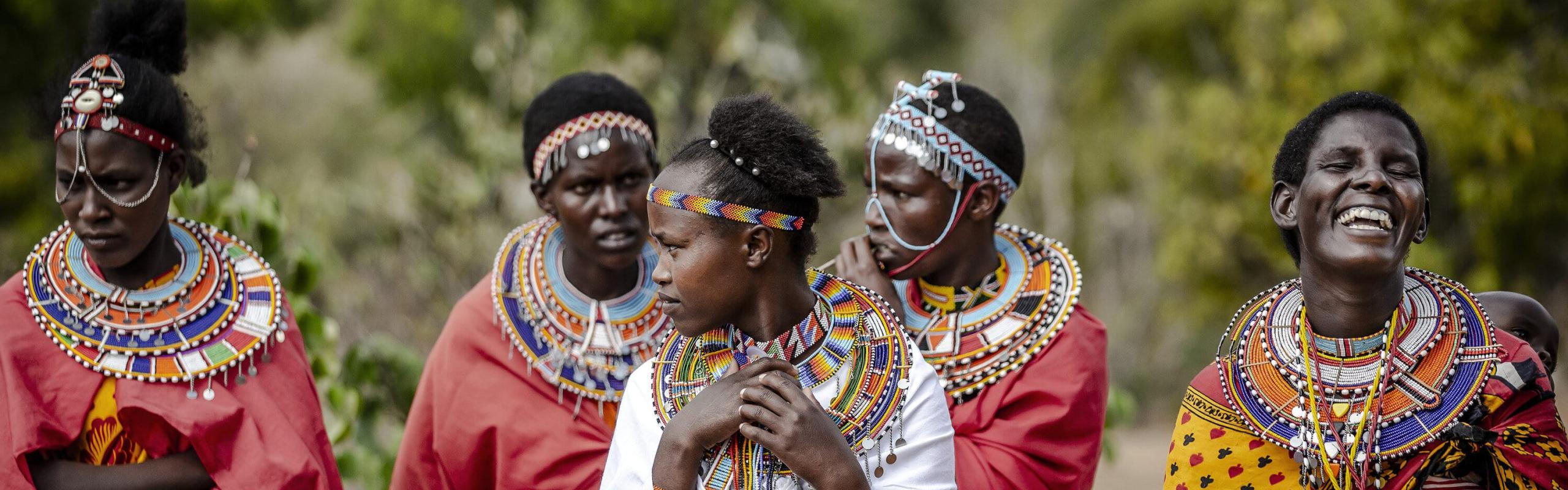 Five members of the Masai community stand together wearing brightly colored traditional clothing and laughing.