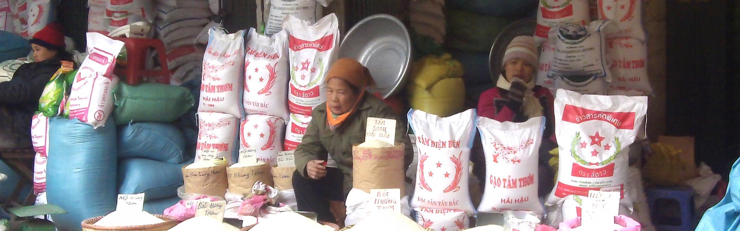 Rice traders sit in outdoor market in Vietnam surrounded by bags of rice.