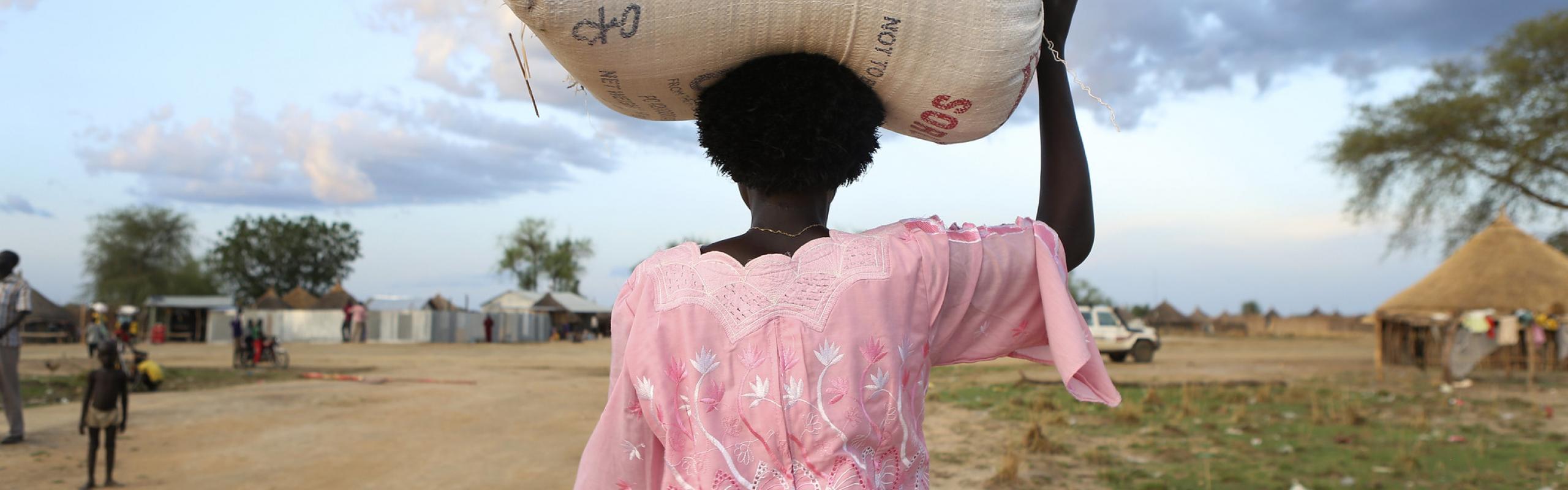 Une femme sud-soudanaise en robe rose s'éloigne de la caméra le long d'un chemin de terre, portant un sac de lentilles et de céréales sur la tête.