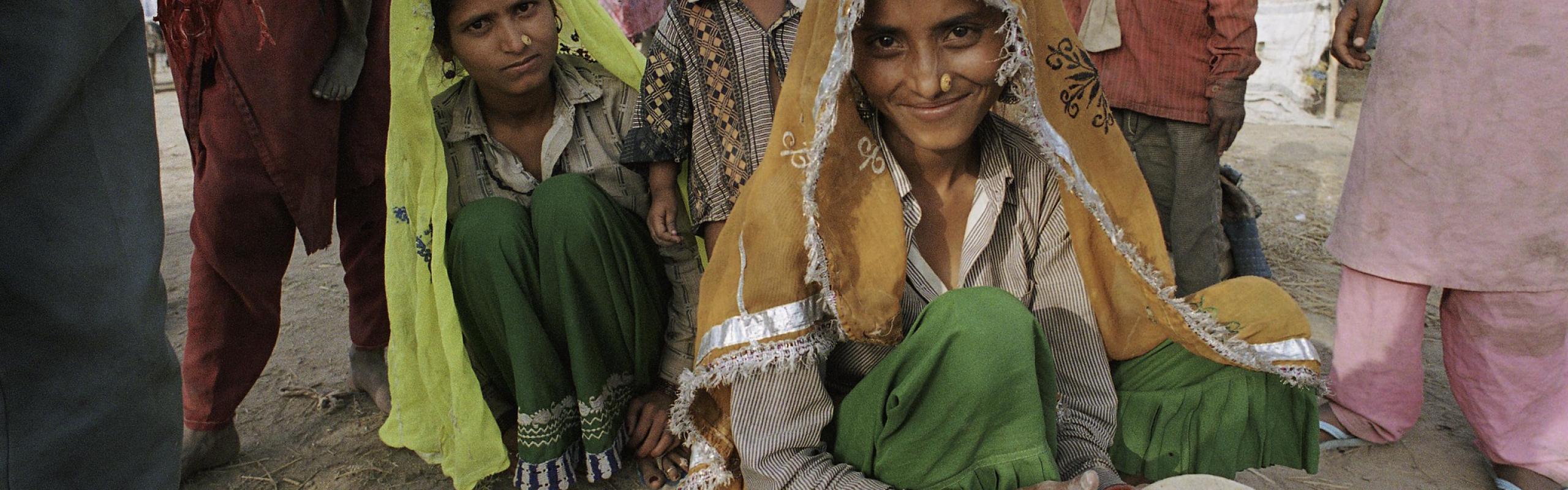 A Bangladeshi woman crouches in front of an outdoor cookstove as she prepares naan. Three children crouch and stand behind her. 