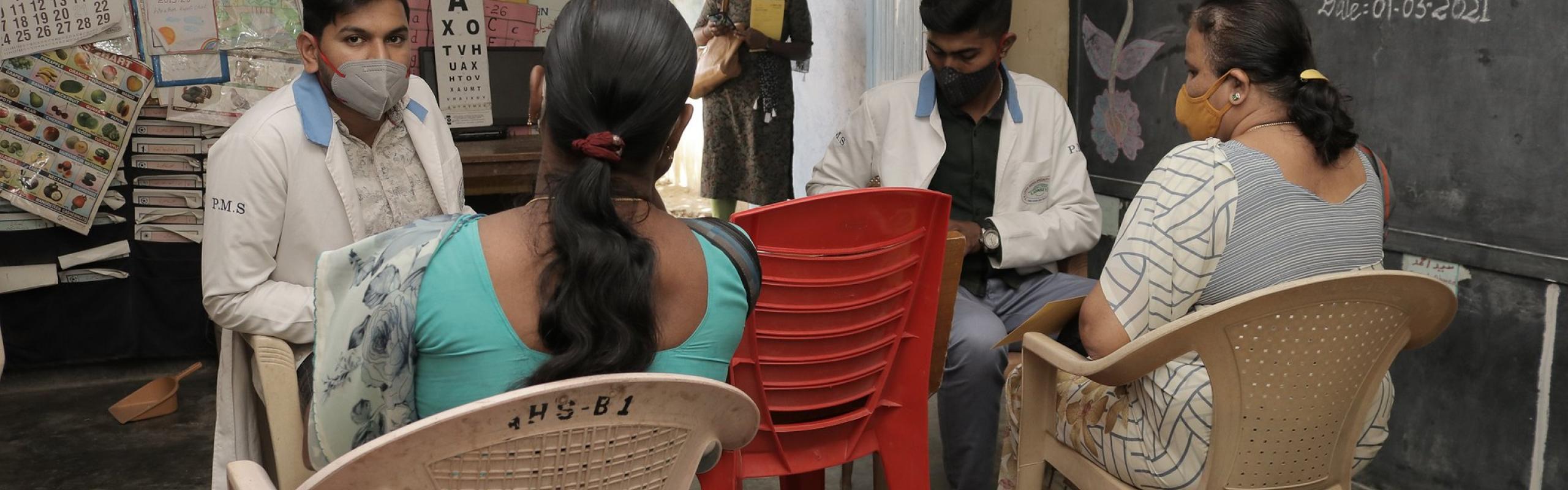 Two women sit in a small room with two male doctors; all wear face masks. The room is part of community health outreach program in Karnataka, India