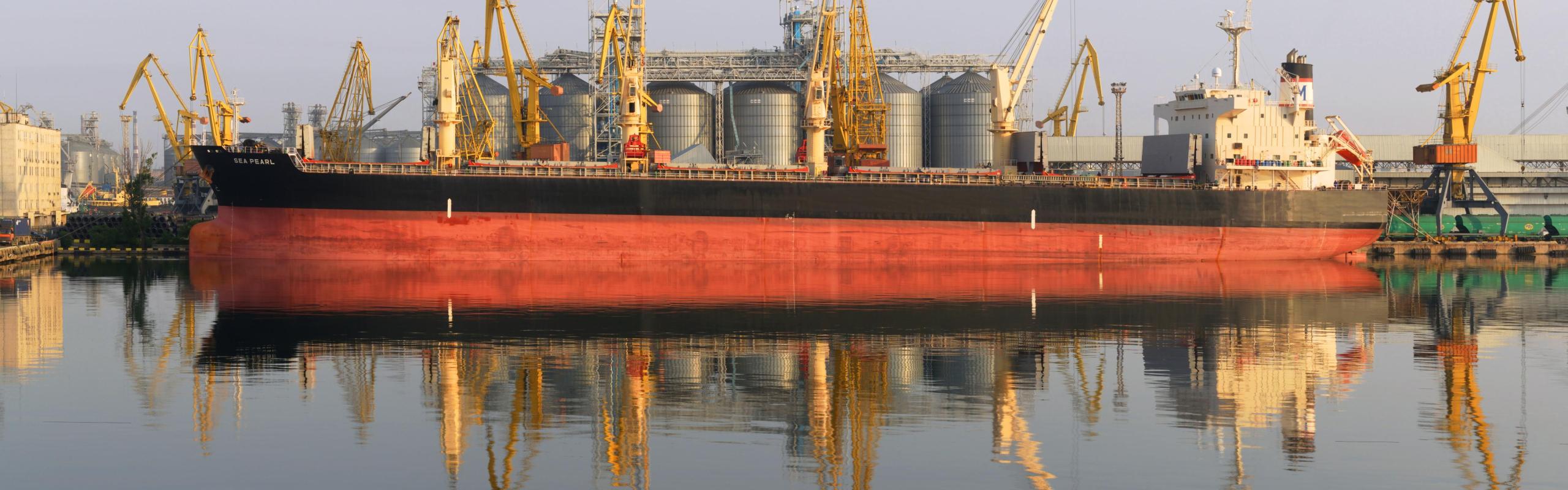 Grain shipping at sea grain terminal. Large red and black carrier ship being loaded with grain in port of Odessa delivering to Brazil. Yellow cranes and silver grain silos are in the background.