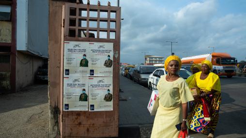 Two women in Lagos, Nigeria carry food and supplies past signs about COVID-19