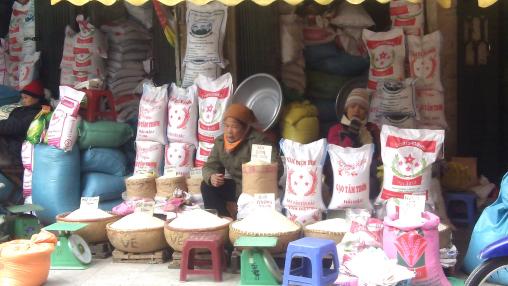 Rice traders sit in outdoor market in Vietnam surrounded by bags of rice.