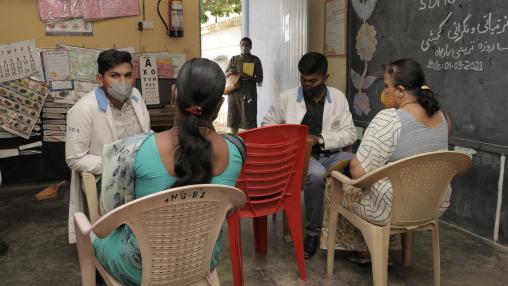 Two women sit in a small room with two male doctors; all wear face masks. The room is part of community health outreach program in Karnataka, India