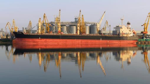Grain shipping at sea grain terminal. Large red and black carrier ship being loaded with grain in port of Odessa delivering to Brazil. Yellow cranes and silver grain silos are in the background.